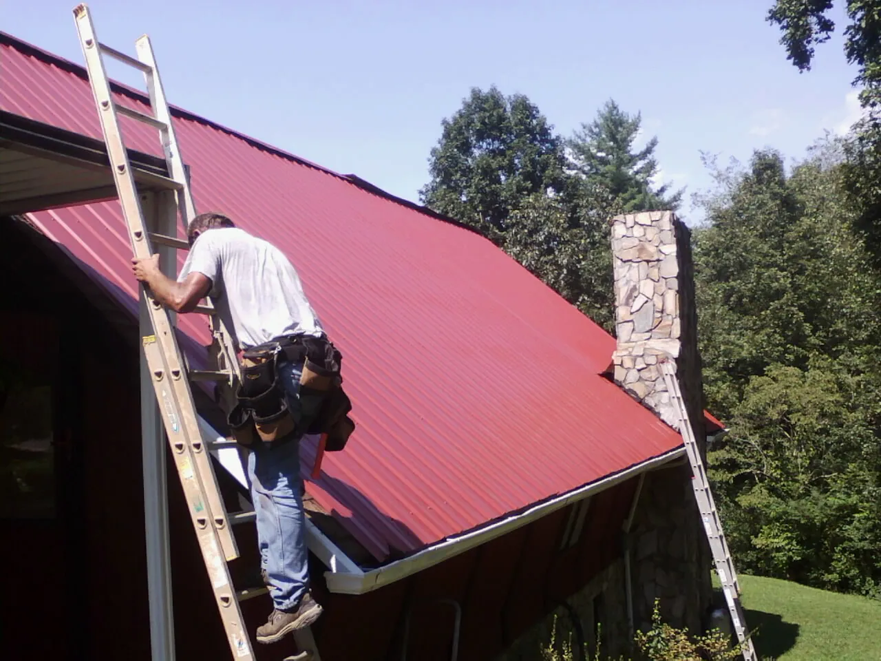 Roofer on a ladder beside a red metal roof and stone chimney — Soffit & Fascia Repair by Roofing Brothers in Point Harbor